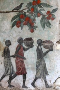 A traditional Indian-style mural painting featuring three figures walking in a line carrying baskets filled with round red fruits, set against a weathered textured wall with a fruiting branch above.